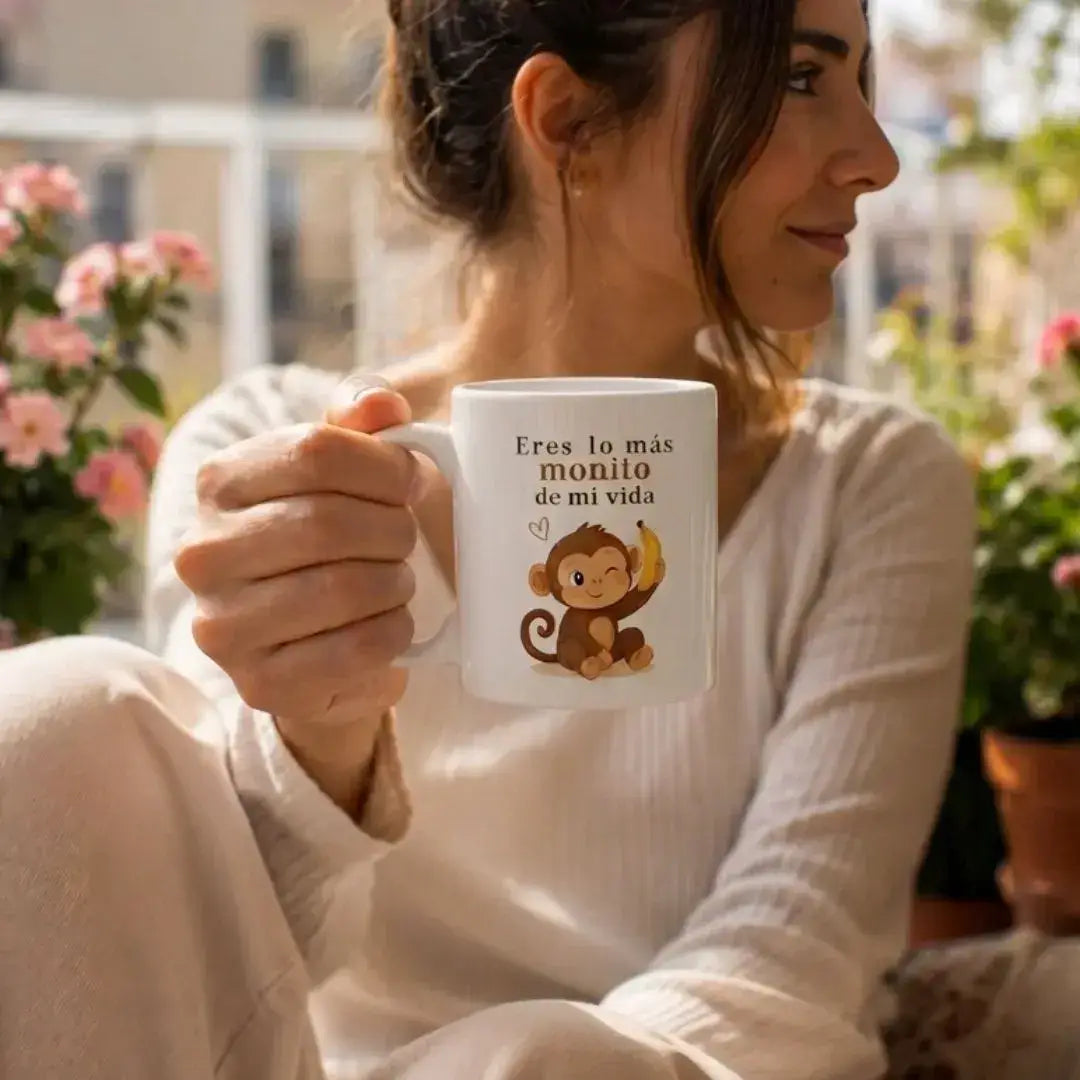 Chica de perfil en una terraza luminosa sosteniendo una taza blanca premium. La taza muestra una ilustración de un tierno monito con un plátano y la frase 'Eres lo más monito de mi vida'. Ambiente acogedor con plantas de fondo.