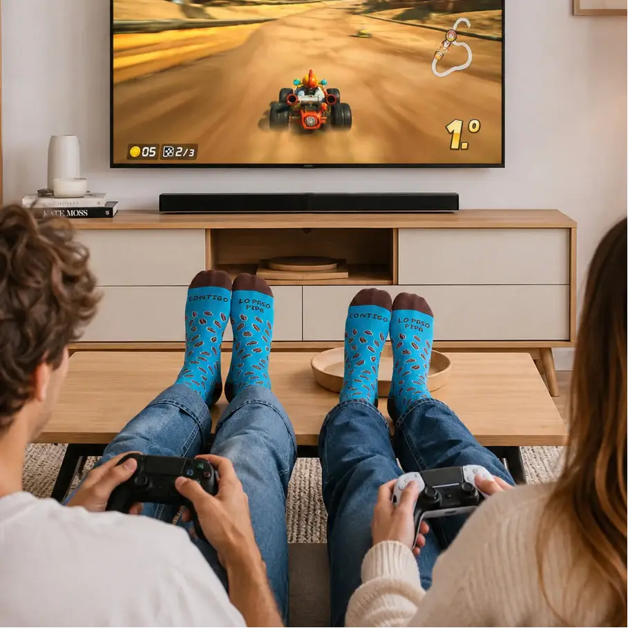 Dos personas de espaldas jugando videojuegos en un salón moderno de colores claros, con los pies apoyados sobre una mesa de centro y llevando calcetines azules con el mensaje 'Contigo lo paso pipa'. Frente a ellos, una gran pantalla muestra un videojuego. Iluminación equilibrada y estilo de catálogo premium