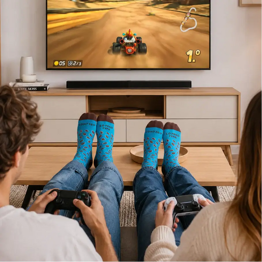Dos personas de espaldas jugando videojuegos en un salón moderno de colores claros, con los pies apoyados sobre una mesa de centro y llevando calcetines azules con el mensaje 'Contigo lo paso pipa'. Frente a ellos, una gran pantalla muestra un videojuego. Iluminación equilibrada y estilo de catálogo premium
