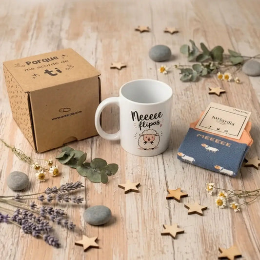 Set de regalo premium con taza blanca ilustrada con una oveja y la frase 'Meeee flipas', caja de cartón kraft y calcetines azules a juego. Decoración minimalista sobre fondo de madera clara con estrellas de madera, piedras de río y hojas de eucalipto con flores blancas bajo iluminación de estudio profesional.