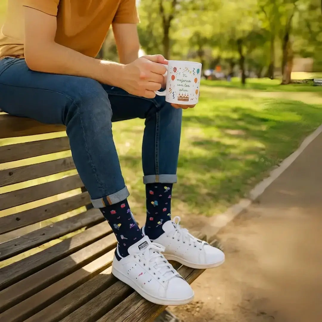 Fotografía de estilo de vida de un hombre sentado en un banco de madera de un parque, sosteniendo una taza blanca con la frase 'Tú mejoras con los años' decorada con globos. Lleva unos calcetines azul marino estampados y unas zapatillas blancas en un fondo natural desenfocado.