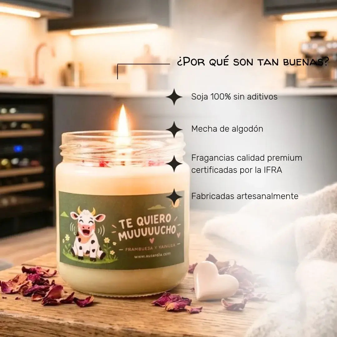 Una vela artesanal de soja con un diseño divertido de vaca que dice "TE QUIERO MUUUUUCHO" en una mesa de madera con pétalos de rosa. La vela tiene una llama encendida y está rodeada de corazones y una toalla. Un cartel con texto en español destaca los beneficios de la vela: soja 100% sin aditivos, mecha de algodón, fragancias de calidad premium certificadas por la IFRA y fabricación artesanal. Un fondo de cocina borroso con un estante para vinos sugiere un ambiente romántico. Esta vela artesanal es un regal